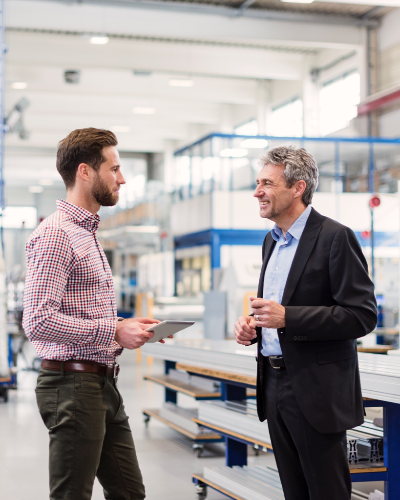 Två män som diskuterar produktionsledning inom industriell tillverkning i en modern fabrik; den ene håller i en surfplatta, medan den andre är klädd i kostym och ler.