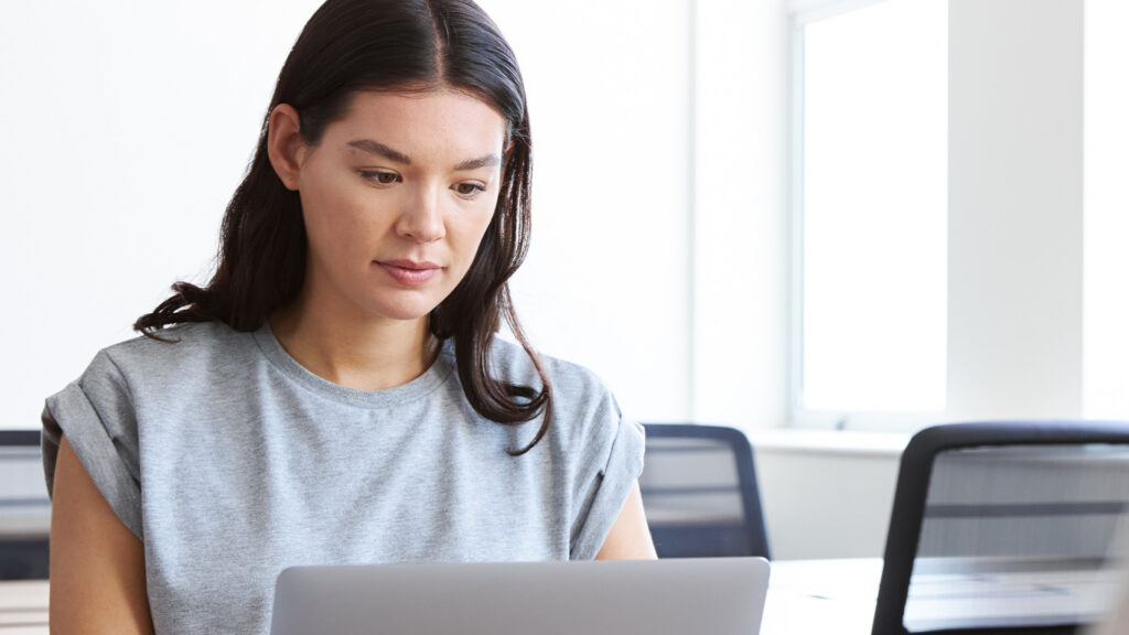 Kvinna med långt mörkt hår i grå t-shirt arbetar på en laptop i ett ljust, modernt kontor och söker efter Urvalsprov för YH - tips & exempel från YH Akademin.