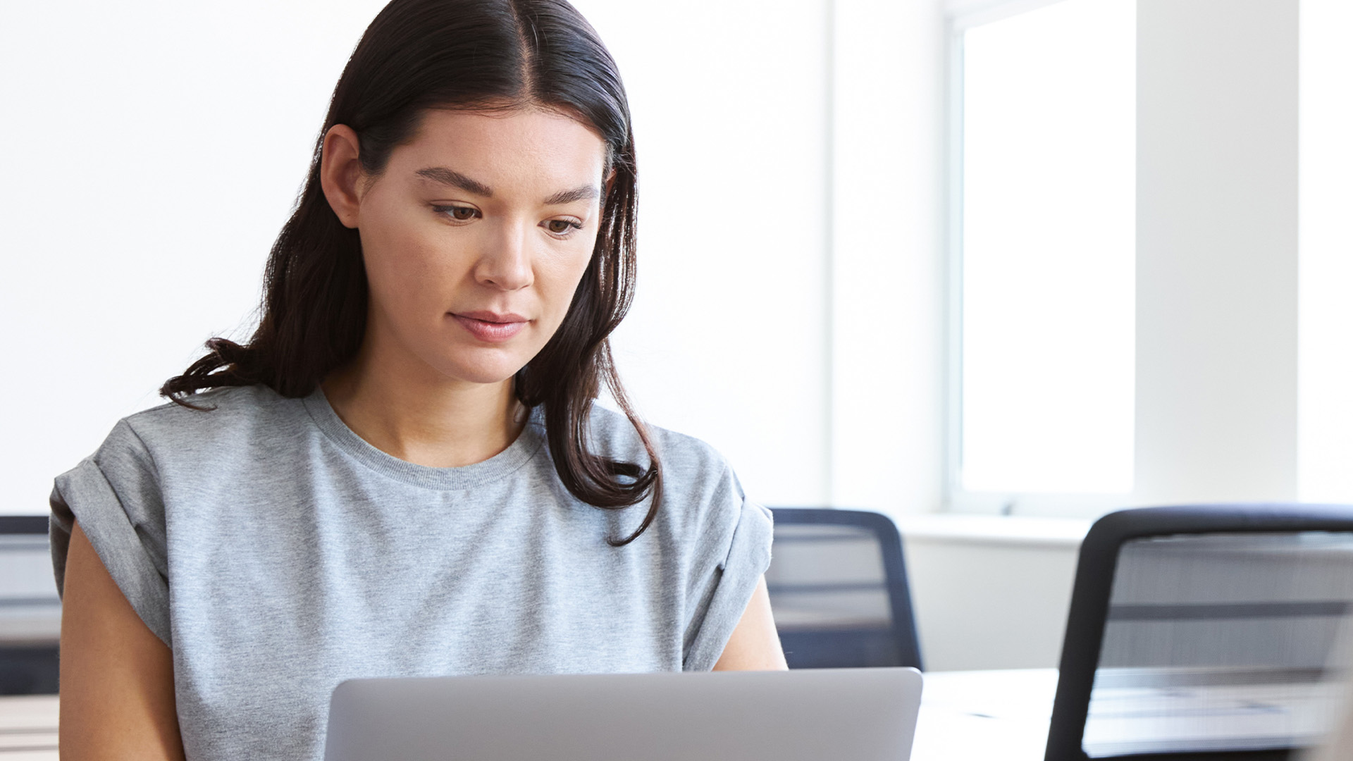 Kvinna med långt mörkt hår i grå t-shirt arbetar på en laptop i ett ljust, modernt kontor och söker efter Urvalsprov för YH - tips & exempel från YH Akademin.