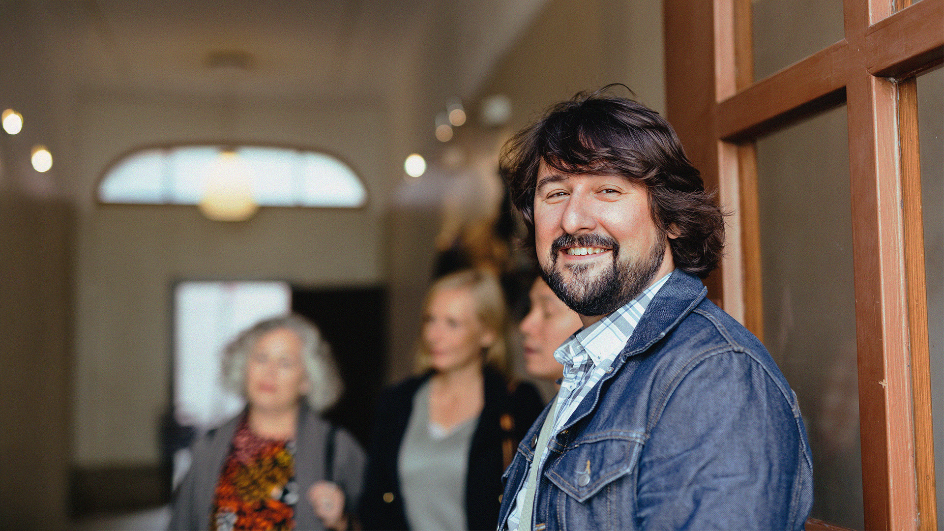 Leende man i jeansjacka står vid en dörr och visar upp sin avslappnade YH-flex-stil, med tre personer suddiga i bakgrunden.