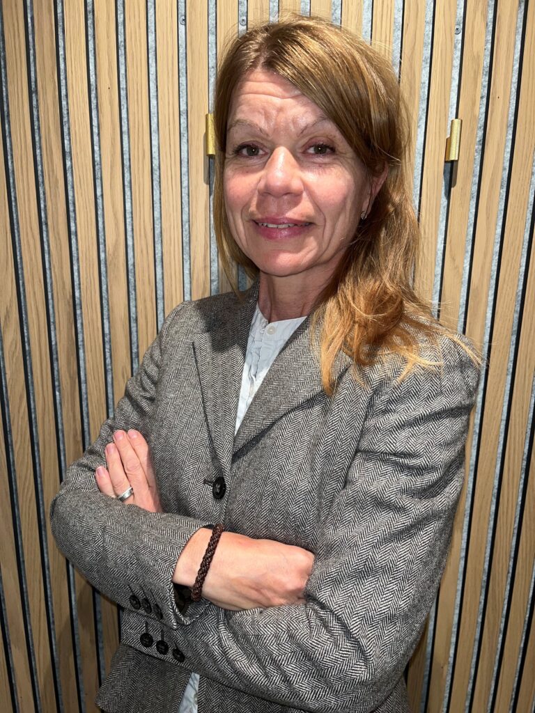 En kvinna i grå kavaj står med armarna i kors och ler framför en träribbvägg som representerar Amphi Tech i ledningsgruppen - Teknisk säljare inom processteknik och vattenhantering.