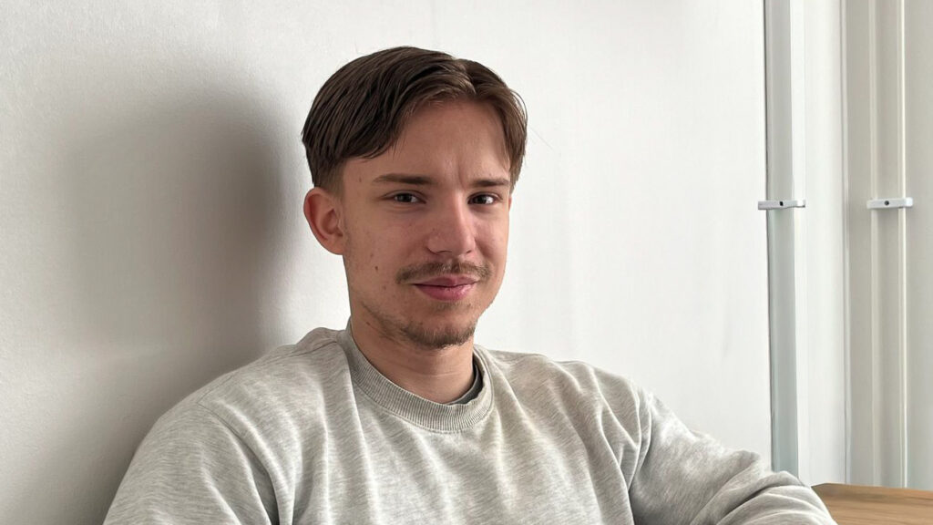 En ung man med kort brunt hår sitter inomhus, klädd i en ljusgrå tröja och med ett mjukt leende - han arbetar som teknisk säljare inom processteknik och vattenhantering i Feralco i ledningsgruppen.