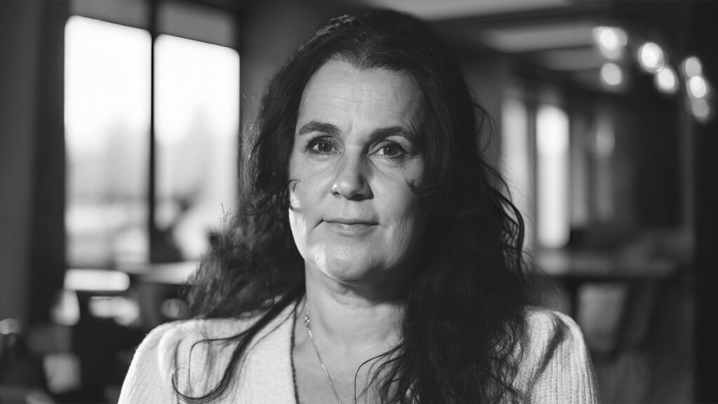 En kvinna med långt mörkt hår tittar in i kameran i en mjukt upplyst inomhusmiljö, som fångar den eftertänksamma atmosfären i YH Akademin i Almedalen.