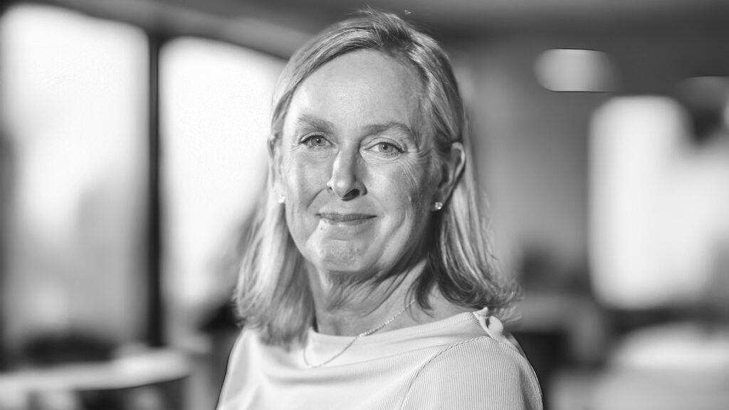 En kvinna med axellångt hår ler mjukt inomhus på YH Akademin i Almedalen, med suddiga fönster och lampor i bakgrunden.