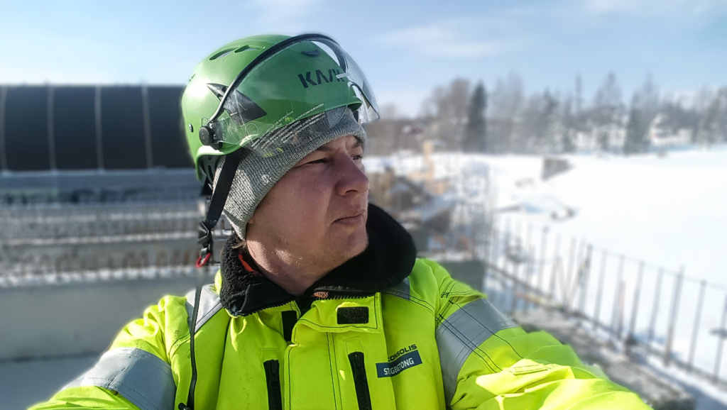 En byggnadsarbetare i grön hjälm och gul jacka står utomhus på en snöig arbetsplats och förkroppsligar andan i 