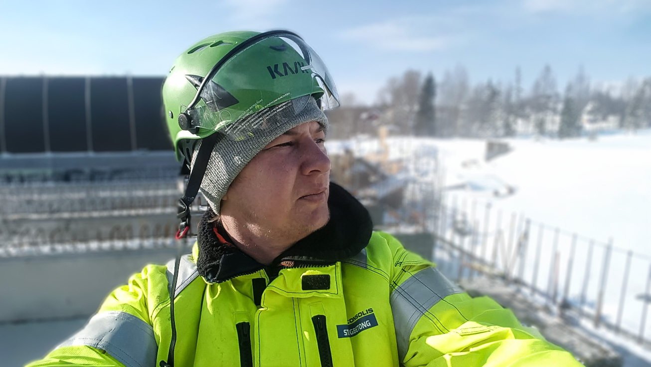 En byggnadsarbetare i grön hjälm och gul jacka står utomhus på en snöig arbetsplats och förkroppsligar andan i 