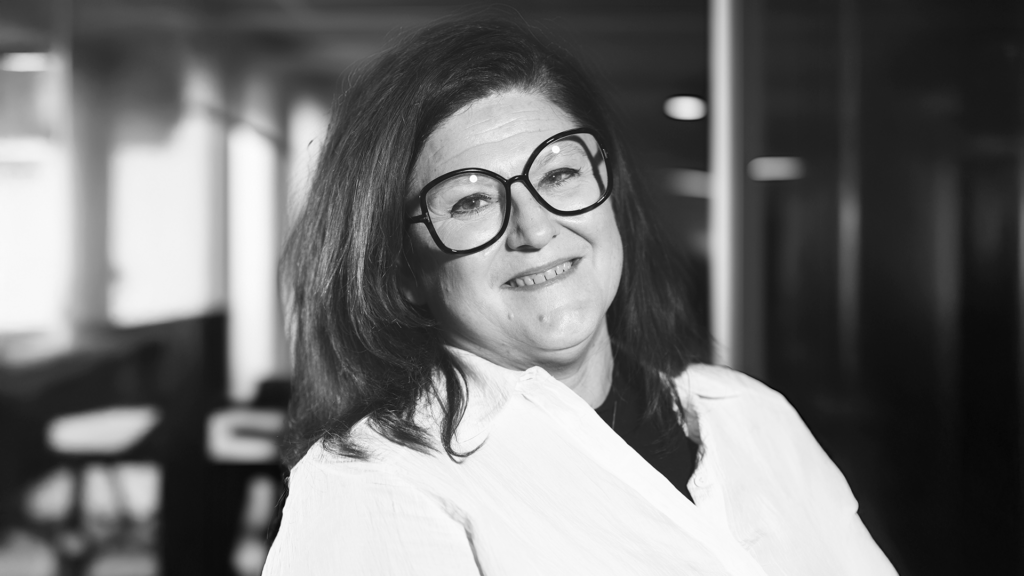 Kvinna med långt mörkt hår, stora glasögon och vit skjorta ler i en inomhusmiljö på YH Akademin i Almedalen.