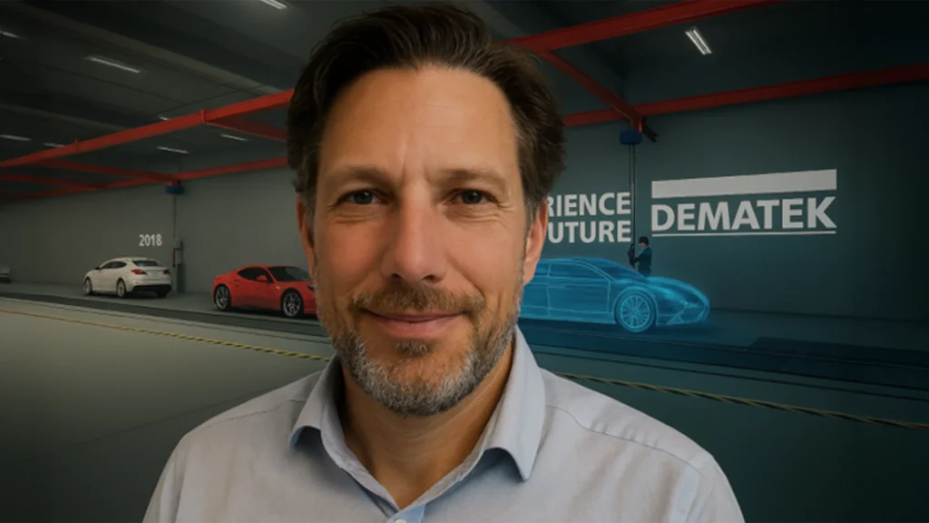 Leende man i skjorta med krage står i en modern bilhall med DEMATEK och bilar i bakgrunden. 