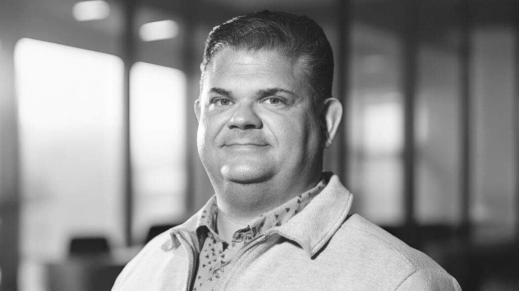 En man i ljus kavaj ler i en kontorsmiljö med suddiga fönster i bakgrunden, föreställande YH Akademin i Almedalen.