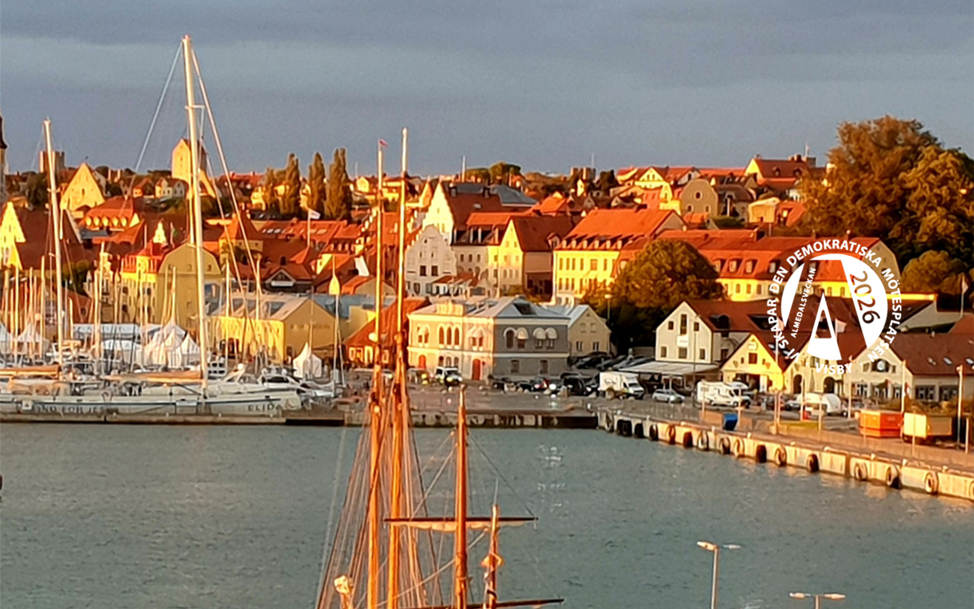 En hamn med segelbåtar, solbelysta byggnader och till höger en logotyp med texten 