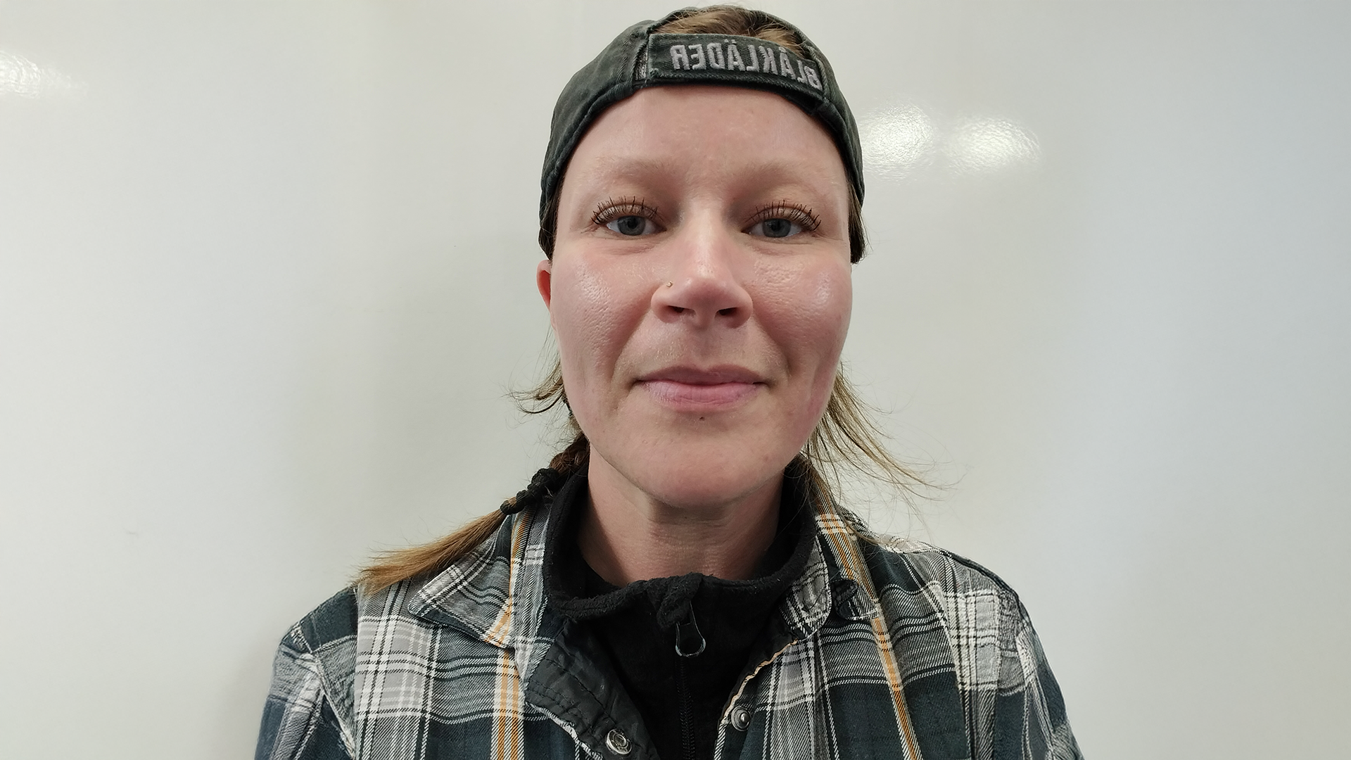 Kvinna i rutig skjorta och bakåtvänd keps, lätt leende framför en vit bakgrund - perfekt för att fånga det avslappnade självförtroendet hos en modern köttförädlare.