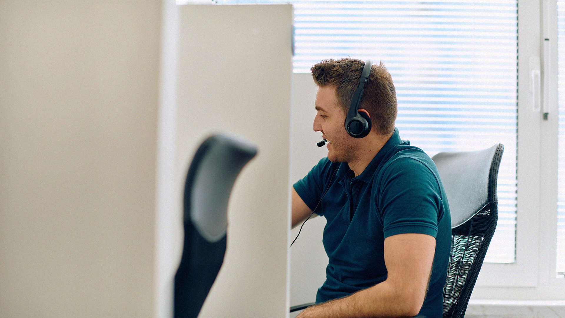 Man med headset ler medan han arbetar vid ett skrivbord i ett ljust kontor med persienner i bakgrunden.