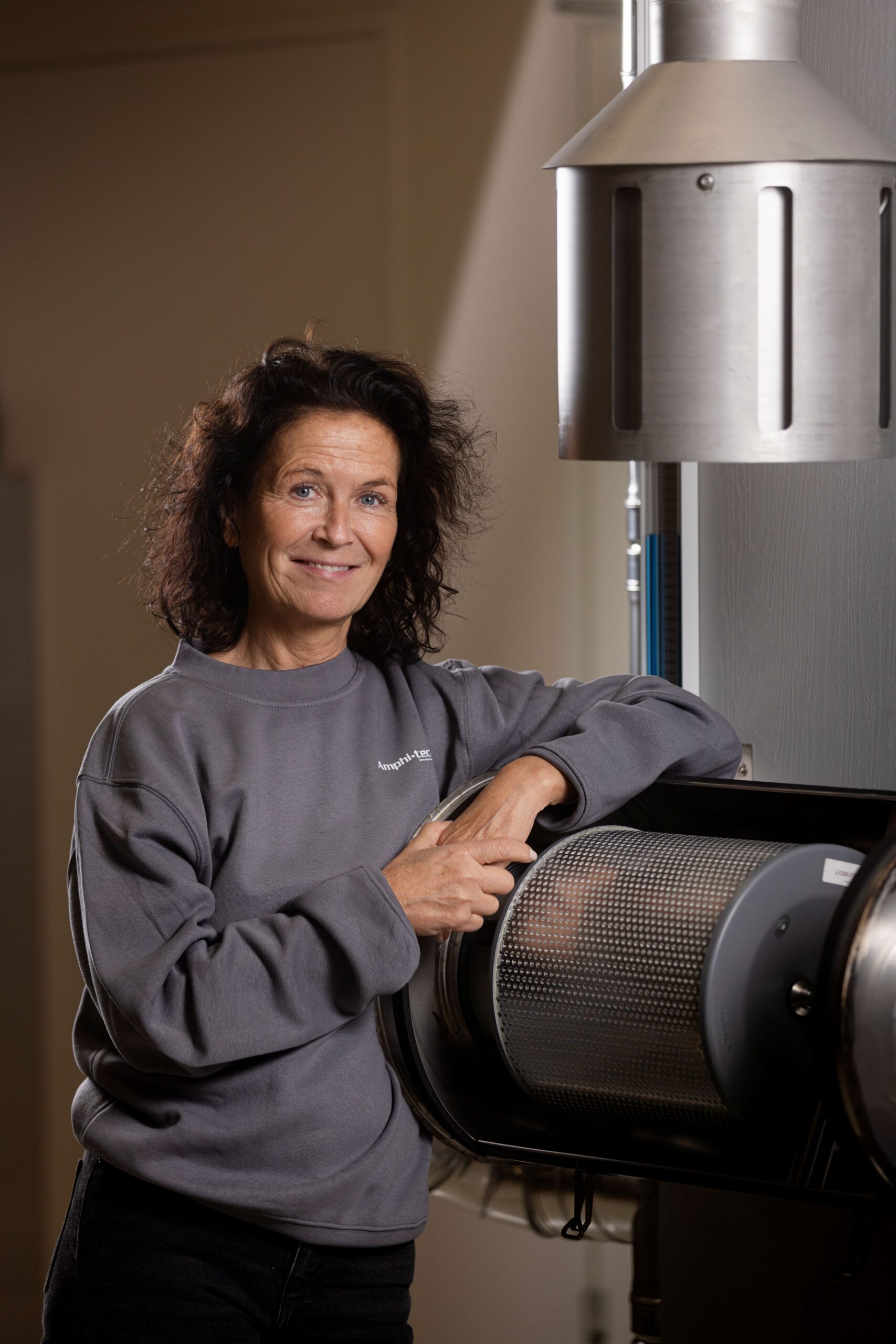 Kvinna med lockigt hår i grå tröja står bredvid industriell metallutrustning och ler mot kameran som representerar Cranemech i ledningsgruppen - Kran- och traverstekniker.