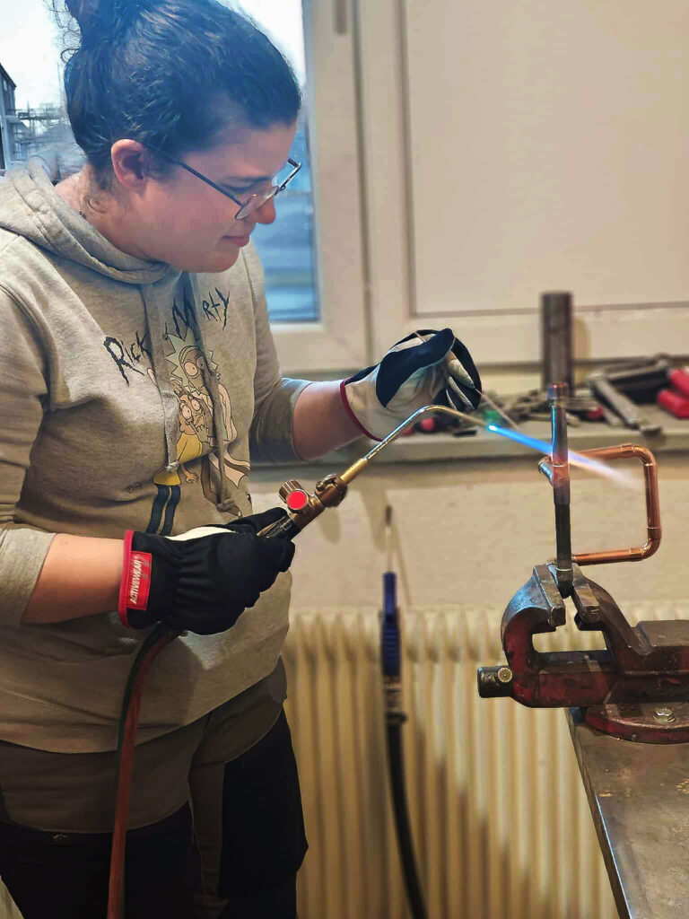 En person, möjligen en köttförädlare, använder en blåslampa på ett metallrör som hålls i ett skruvstäd, iklädd handskar och skyddsglasögon.