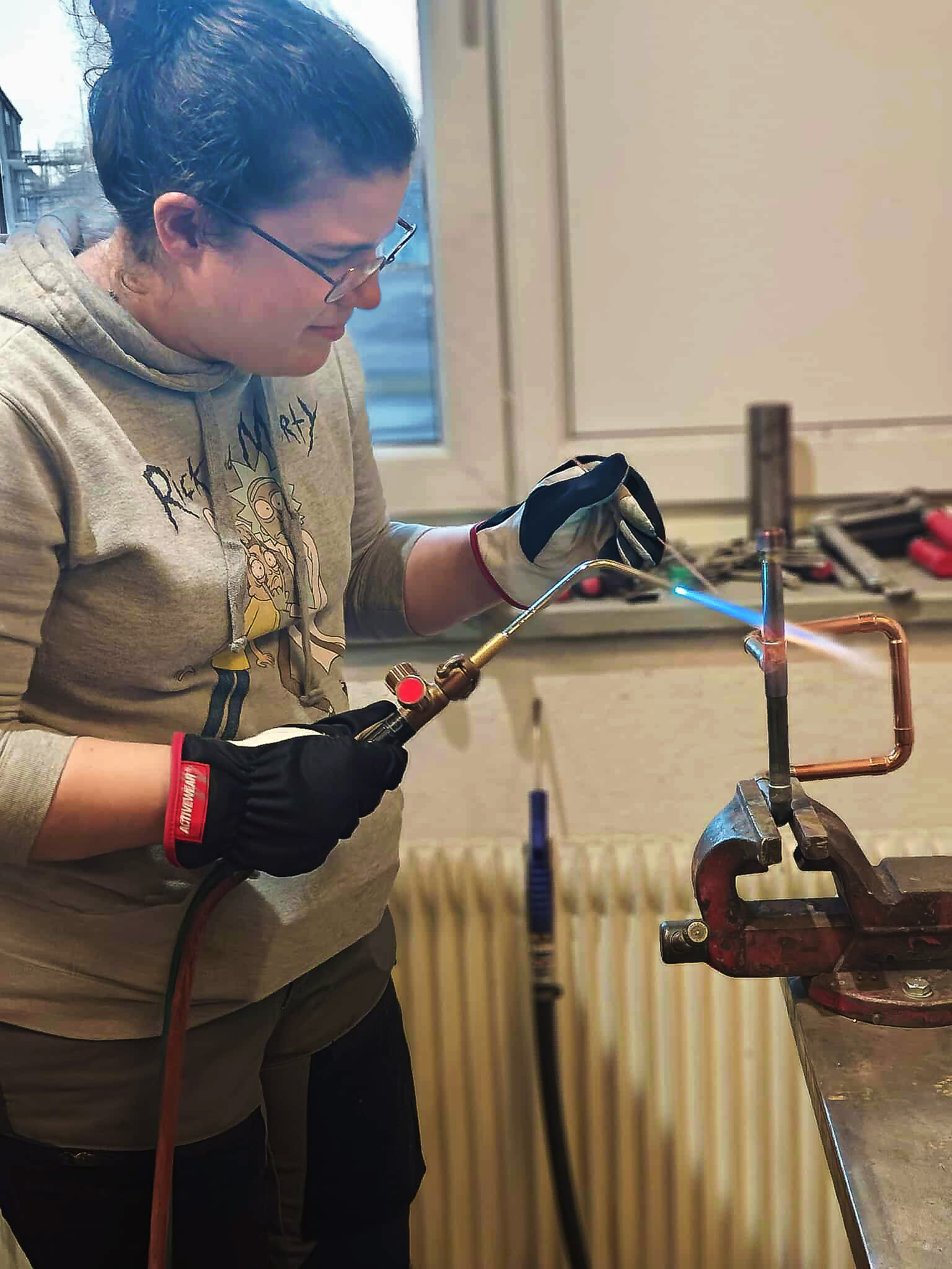 En underhållsmekaniker använder en blåslampa för att bearbeta metall som hålls i ett skruvstäd, med handskar och skyddsglasögon som skydd.