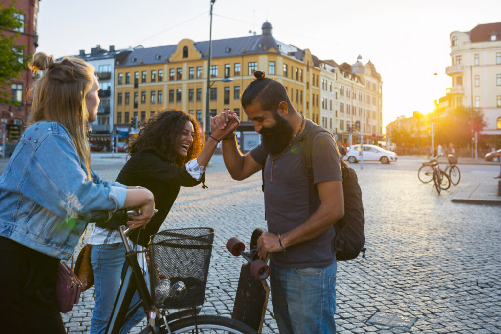 Tre vänner hälsar glatt på varandra på ett torg i solnedgången, med cyklar och en skateboard i blickfånget.