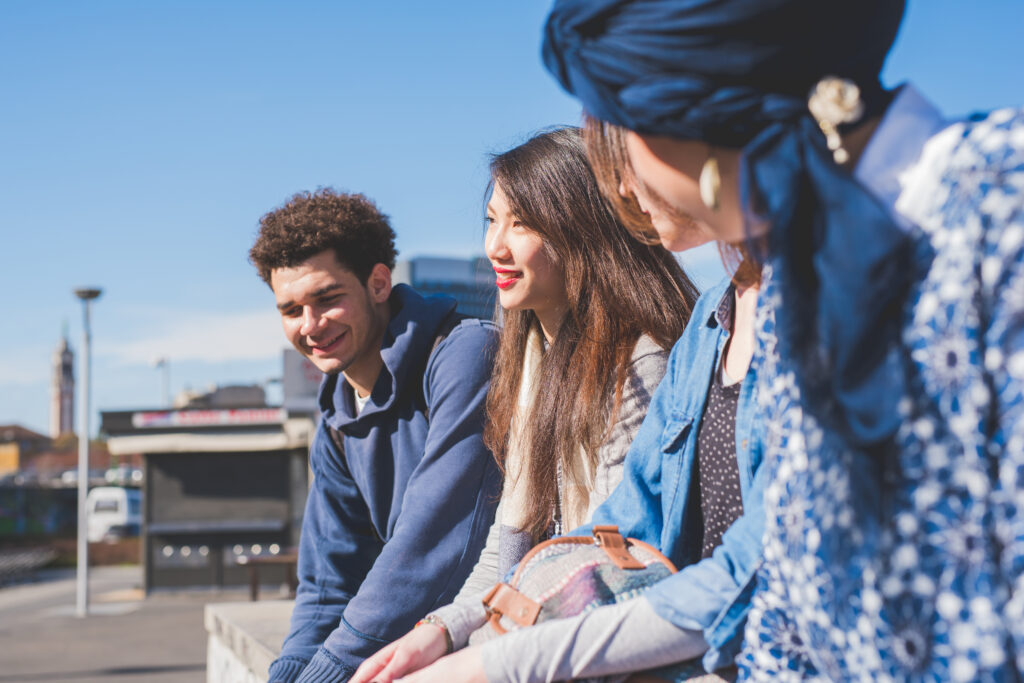 Tre unga vuxna pratar och ler utomhus en solig dag, med en blå himmel i bakgrunden, och diskuterar fördelarna med Stipendium inom yrkeshögskolan - YH Akademin.