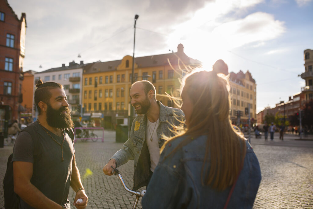 Tre vänner skrattar och pratar utomhus en solig dag, med färgglada byggnader i bakgrunden, och delar historier om sina erfarenheter av Stipendium inom yrkeshögskolan - YH Akademin.