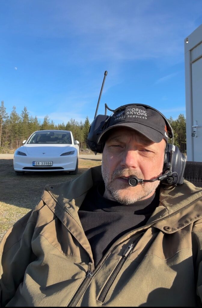 En man med headset och keps sitter utomhus; en vit Tesla står parkerad bakom honom en solig dag – vilket illustrerar den moderna utbildningsupplevelsen vid YH Akademin – Nationella Yrkesutbildning för blivande underhållsmekaniker.