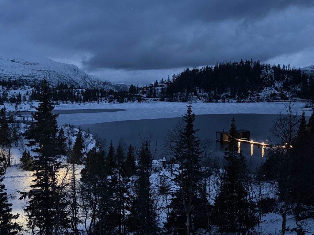 En snötäckt sjö med tallar, en brygga med belysning och berg under en molnig kvällshimmel – en bild som fångar den fridfulla stämningen nära YH Akademin – Nationella Yrkesutbildning – Underhållsmekaniker.