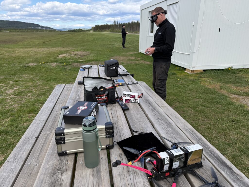 En person med FPV-glasögon styr en drönare utomhus, med utrustningen uppställd på ett bord i närheten – vilket visar de praktiska färdigheter som lärts ut vid YH Akademin – Nationella Yrkesutbildning för blivande underhållsmekaniker.