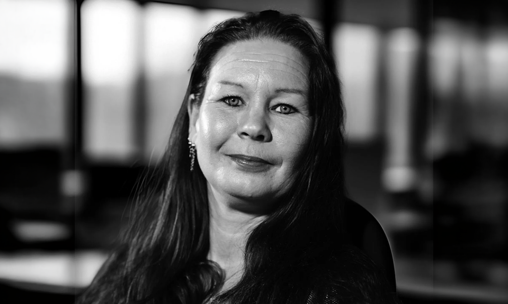 En kvinna med långt mörkt hår som ler svagt och bär örhängen, fotograferad mot en enfärgad bakgrund i svartvitt. Ett porträtt för YH Akademin.