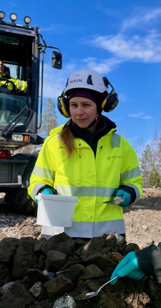 En kvinna i skyddsutrustning håller en vit behållare på en stenig plats; i bakgrunden syns maskiner och en blå himmel, vilket symboliserar resan från vården till miljöutredning: ”Det bästa yrkesvalet jag har gjort.”.