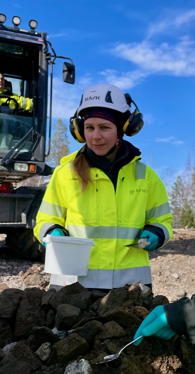 En kvinna i skyddsutrustning håller en vit behållare på en stenig plats; i bakgrunden syns maskiner och en blå himmel, vilket symboliserar resan från vården till miljöutredning: ”Det bästa yrkesvalet jag har gjort.”.