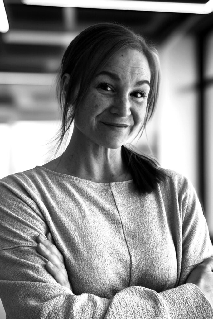 En kvinna med hästsvans ler med armarna i kors, stående inomhus i ett modernt, väl upplyst rum. Svartvitt.