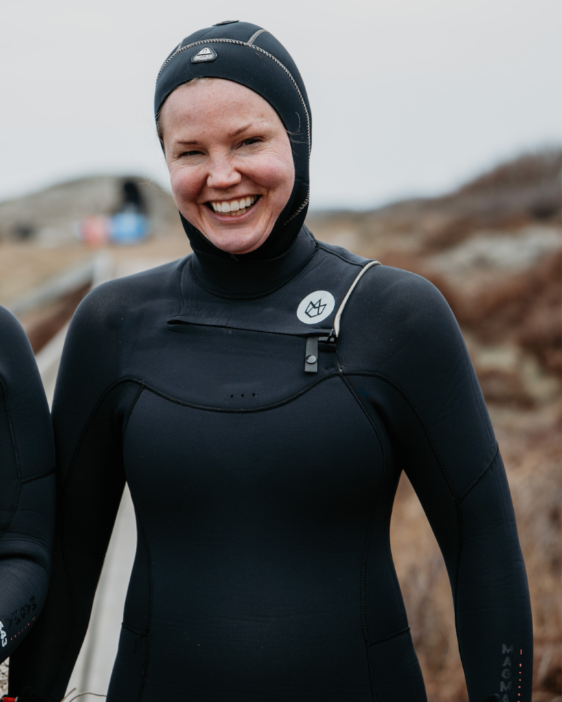 En leende person i svart våtdräkt och huva, som står utomhus nära vattnet med en suddig naturbakgrund – en bild som för tankarna till resan från kryddfälten i Skåne till tången i Bohuslän.