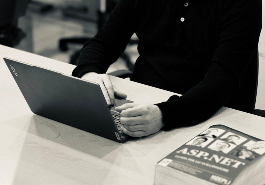 En man som arbetar på en bärbar dator vid ett skrivbord med en ASP.NET-bok i ett datalabb – en bild som fångar resan från ”Från karriärbyte till Rom – så tog Erasmus+ Christoffer till Italien” i detta slående svartvita foto.