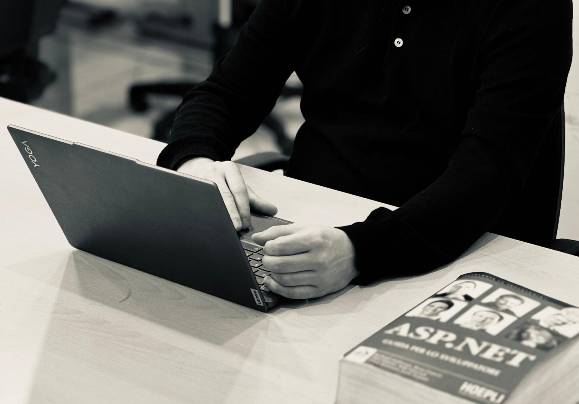 En man som arbetar på en bärbar dator vid ett skrivbord med en ASP.NET-bok i ett datalabb – en bild som fångar resan från ”Från karriärbyte till Rom – så tog Erasmus+ Christoffer till Italien” i detta slående svartvita foto.