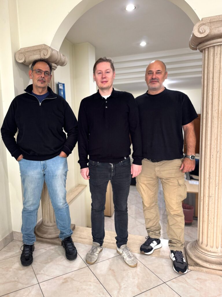 Tre män står tillsammans inomhus mellan två pelare, alla klädda i mörka tröjor och vardagsbyxor – kanske utbyter de berättelser om allt från kryddfält i Skåne till tång i Bohuslän.