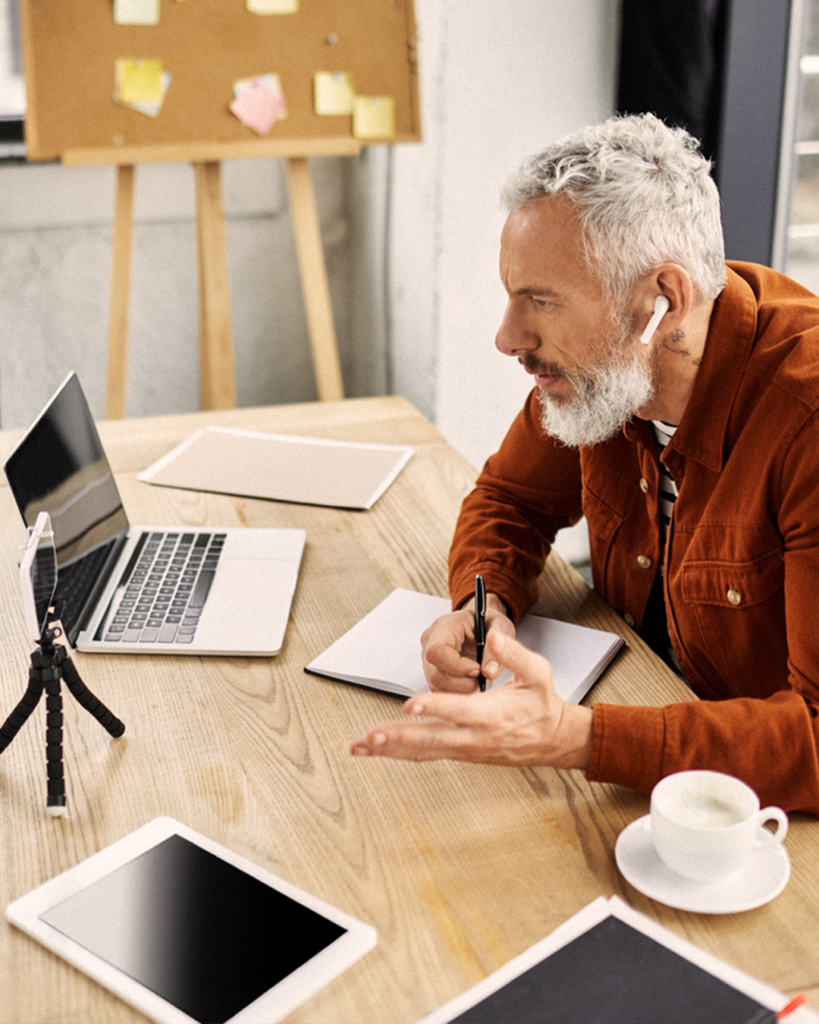 En man med grått hår under ett videosamtal, sittande vid ett skrivbord med en bärbar dator, en anteckningsbok, en surfplatta och en kopp kaffe – deltar i ett virtuellt möte med YH Akademin.